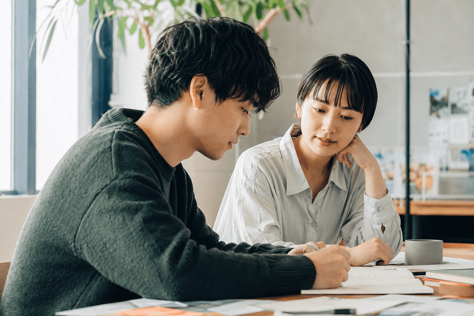 AI生成の撮影イメージ：机を囲み、ノートを見ながら仕事に向き合う二人。実在の社員ではありません。